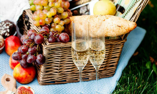 Picnic at the park on a grass, delicious food: basket, wine, grapes, peaches, baguette, cupcakes, figs, cheese, blue tablecloth, wineglass with champagne